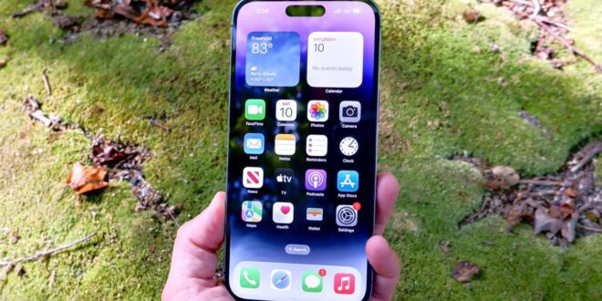 من iPhoneIslam.com، يد تمسك بجهاز iPhone يعرض أيقونات تطبيقات متنوعة مع أداة الطقس على الشاشة الرئيسية، على خلفية مطحونة.