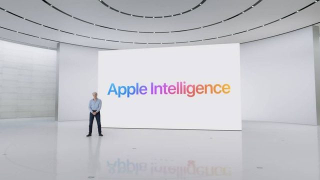 من iPhoneIslam.com، يقف فرد في غرفة بيضاء حديثة بها شاشة كبيرة تعرض كلمة "Apple Intelligence" بأحرف ملونة، ويتابع آخر الأخبار.