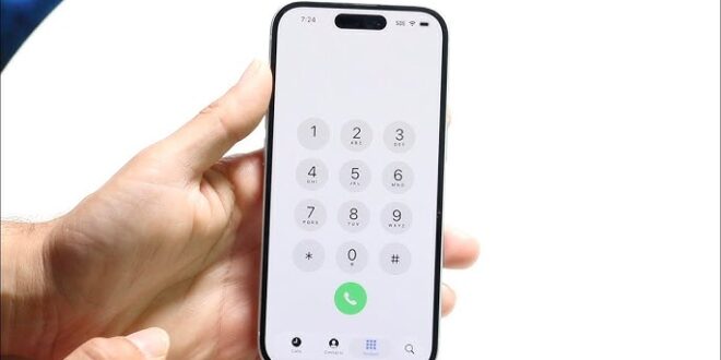 من موقع فون إسلام: يد تحمل هاتفًا ذكيًا يعرض لوحة الاتصال الهاتفي التي تحتوي على لوحة الاتصال الهاتفي مع الأرقام 0-9 والنجمة ومفاتيح الجنيه وزر الاتصال الأخضر - تظهر الميزات الجديدة بعد تحديث iOS 26.